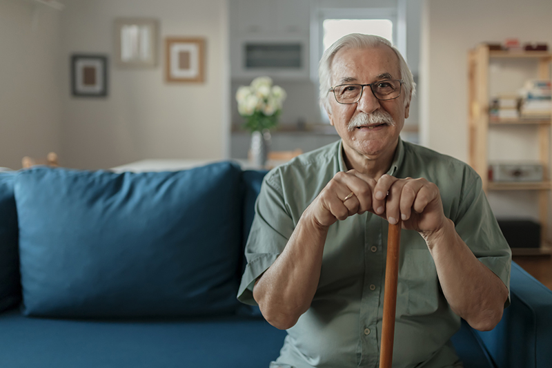 Portrait of happy senior man smiling at home while holding walking cane. Old man relaxing on sofa and looking at camera. Portrait of elderly man enjoying retirement."n